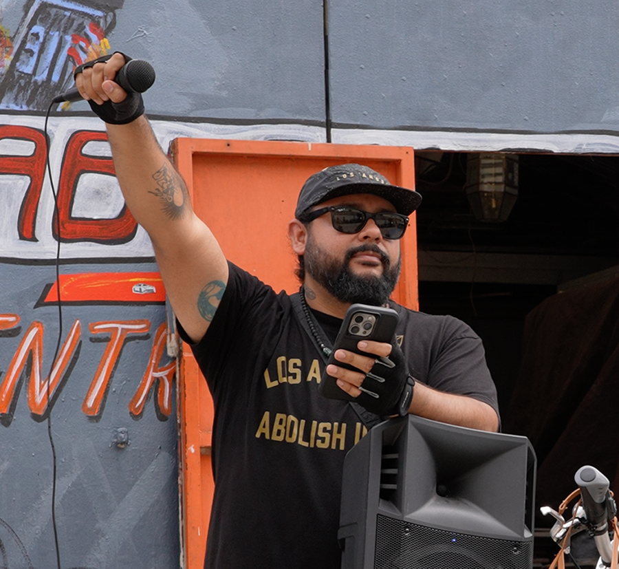 mark speaking at an outdoor event, holds a microphone in his raised hand.